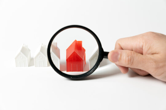 A hand holds a magnifying glass over a single red miniature house model in a row of white plastic houses. Concept for real estate search, property inspection, house hunting