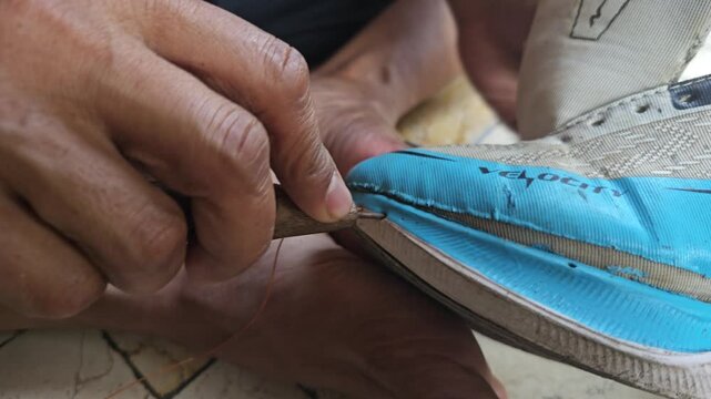 Detailed view of a traditional shoemaker hands using a sharp awl and thread to mend the sole of a blue sneaker utilizing their feet for grip