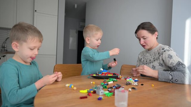 Young mother helps two boys assemble colorful building blocks in kitchen. Cozy family scene showing teamwork, imagination, fine motor skills, and early child development