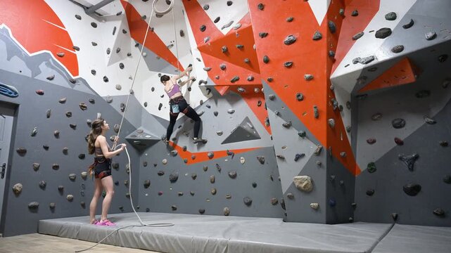 Female climber practicing difficult move in indoor bouldering center with partner nearby. Dynamic footage for determination, skill development, teamwork, and active lifestyle themes