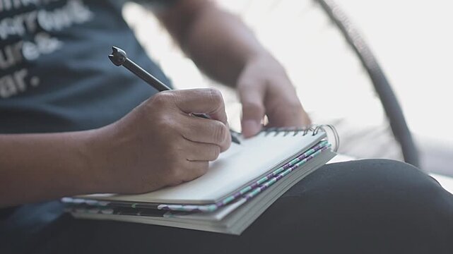 Close up male hands holding pen and writing text in spiral notebook outside. Young man author taking creative notes, planning lifestyle or studying in nature background