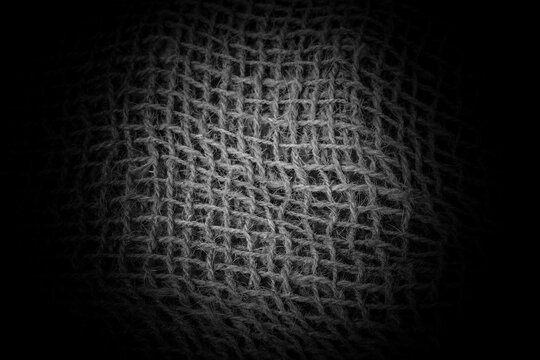 Close-up black and white texture of woven fabric or burlap, featuring a central spotlight effect that fades into darkness at the edges.