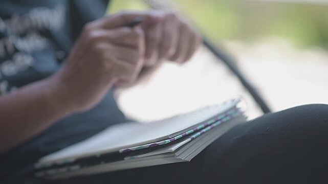 Close up of young man hands holding pen and writing text on blank paper notebook outdoor. Female taking notes, planning, studying or writing a diary in nature background during day