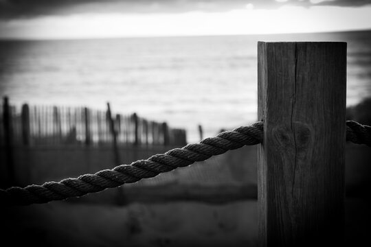 Black and white photograph focusing on a weathered wooden post and a thick, twisted rope railing passing through it, set against the blurred background of a beach in Labenne-Oc&eacute;an. The scene has a dra