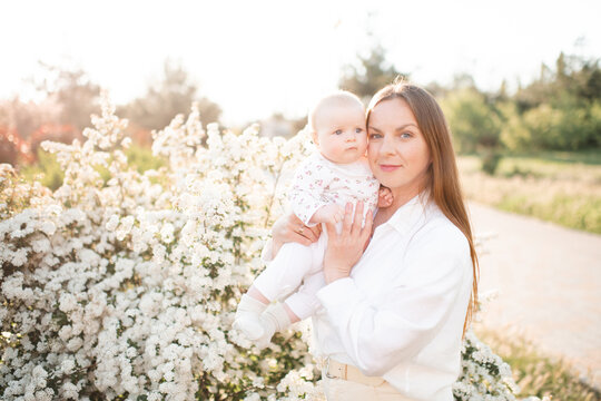 Happy young woman 30-35 year old holding baby over flowers in sunny day outdoors. Summer vacation season. Motherhood. Looking at camera.