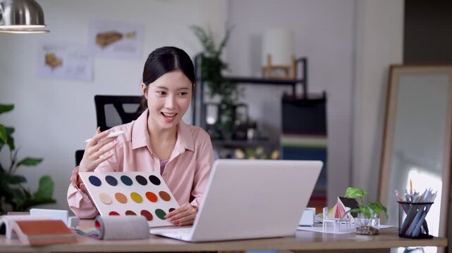 Young Asian interior designer presenting color palette to camera while working on furniture design project with laptop in bright modern home office studio