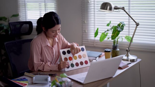 Young Asian furniture designer presenting color palette swatches to camera while working on laptop in bright modern home office studio with natural light
