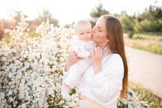 Happy young woman 30-35 year old holding and kissing baby over flowers in sunny day outdoors. Summer vacation season. Motherhood.