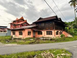 Obraz premium Traditional Malay wooden house with zinc roof in rural village under cloudy sky. Classic Southeast Asian architecture, kampung style home along roadside.