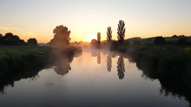 Tranquil river at sunrise with mist rising and trees reflected in the calm water.