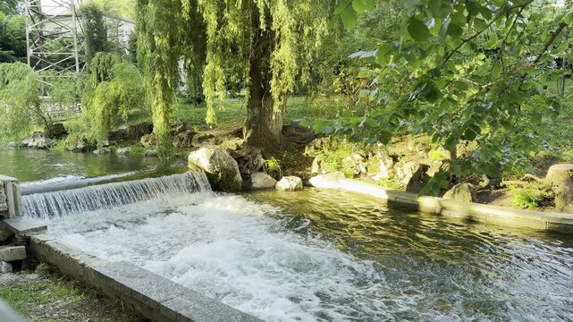a lively waterfall flowing into a rushing park river. This sunny scene brings a joyful and refreshing feeling of peaceful nature.