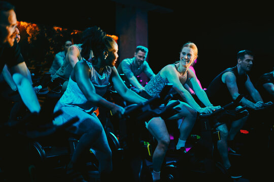 Diverse group enjoying a high-energy spin class in an indoor cycling fitness studio. The room is filled with blue light