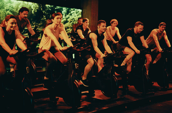 Energetic spin class with diverse adults doing indoor cycling workout in a dim studio