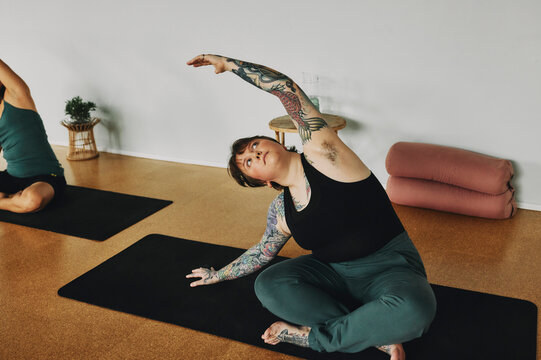 Body positive young woman with tattoos practicing her yoga poses on an exercise mat during a class in a studio. She has armpit hair and short hair
