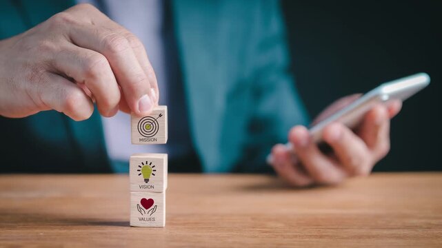 Close-up of a businessman stacking wooden blocks with mission, vision, and values icons, holding a smartphone for corporate strategy, brand philosophy, and company identity planning concept.