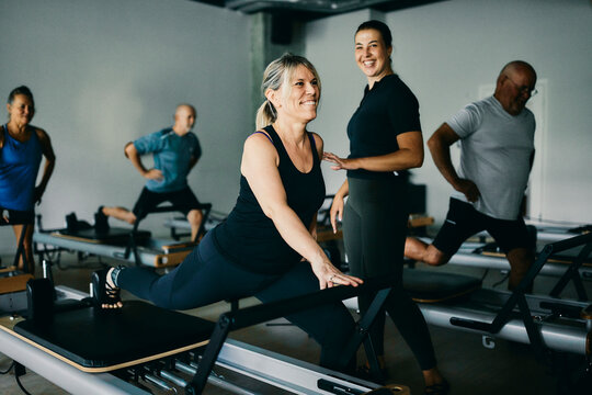 Smiling fitness instructor working with a mature female client during a reformer class with a group of people in a pilates gym