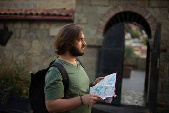 Horizontal travel photo. White young adult man with beard, bob haircut, green t-shirt stands, black backpack walks on old city street, holds paper map, looks at sight. Tourism, road trip, walk concept