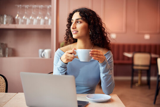 Entrepreneur woman in a cozy cafe with laptop and coffee, enjoying a calm moment today. She sits smiling and relaxed, looking out the window