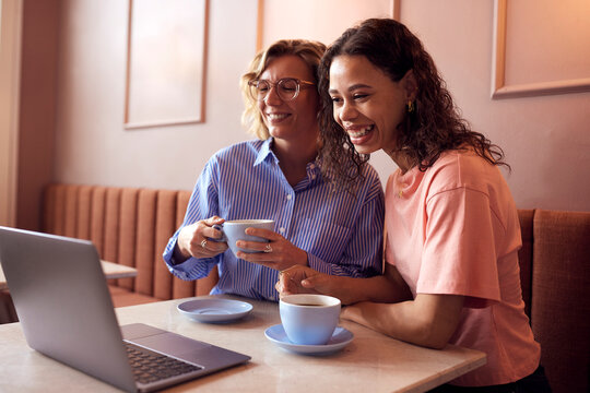 Two multicultural smiling friends in a cozy cafe share a smile over coffee and a laptop, enjoying a casual moment together