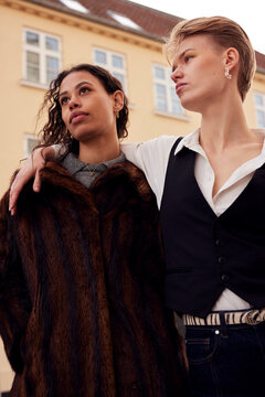 Two diverse friends stand close together in stylish outfits outside a sunny street scene. One has their arm around the other's shoulder