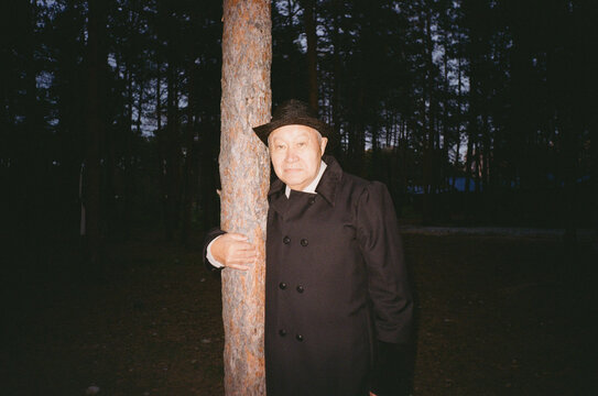 old-aged man in a long black coat leaning against tree
