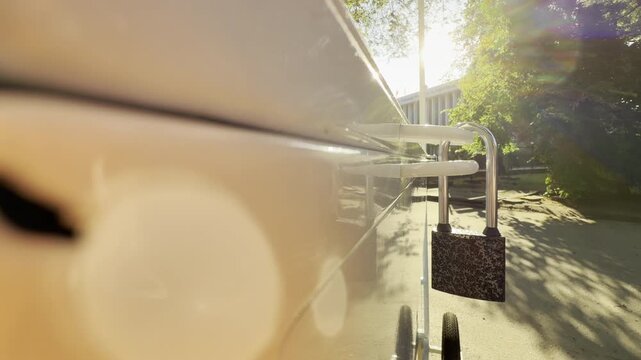 A white ice cream freezer remains securely locked with a padlock. It stands quietly on a sun-drenched park path, evoking a warm, peaceful feeling of morning anticipation.