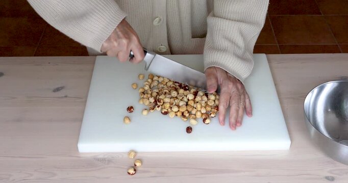 Female hands finely chopping roasted hazelnuts with knife on white cutting board during preparation of traditional Italian meringue cookies. 