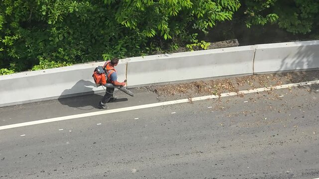 Maintenance worker using a backpack leaf blower to clear leaves and debris from the asphalt edge beside a concrete barrier, supporting highway safety and roadside upkeep