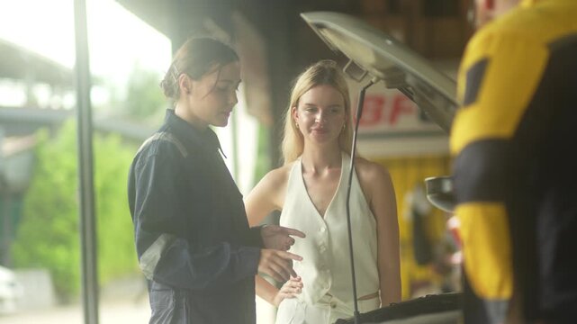 female technician in a dark uniform talks and points at the car engine, consulting a blonde female driver who smiles with confidence under an open car hood. This authentic stock imagery represents exc