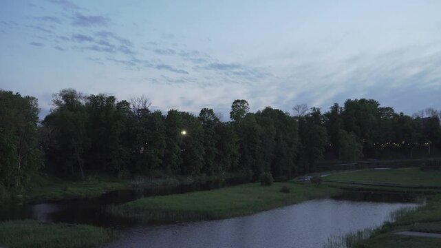 Distant lanterns glow through dark trees by a quiet park pond under a cloudy twilight sky. Ideal for nature documentaries or serene cinematic transition scenes. 