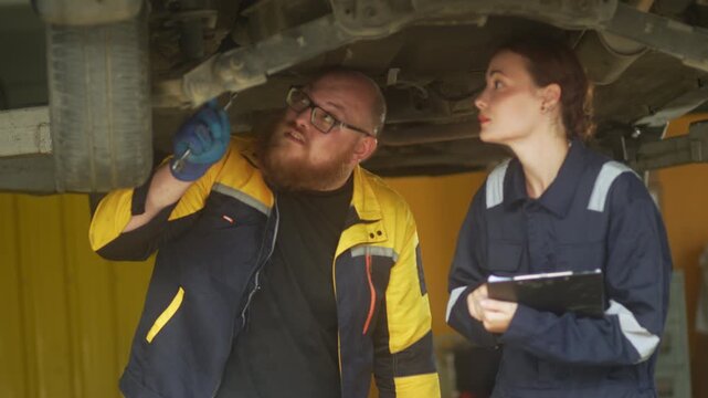 Caucasian male mechanic with glasses and a beard works alongside a female technician, reviewing a digital tablet and giving a thumbs-up. This high-energy commercial imagery represents gender diversity