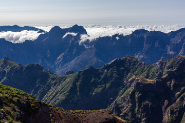 Obraz premium Volcanic peaks dominate Madeira's highland landscape. Clouds drift over rugged cliffs and lush green valleys. Panoramic view from Pico do Areeiro showcases Atlantic island scenery.