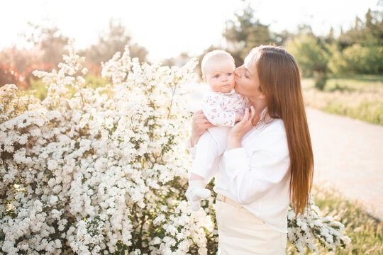 Happy young woman 30-35 year old holding and kissing baby over flowers in sunny day outdoors. Summer vacation season. Motherhood.