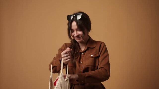 Stylish woman putting fresh vegetables and fruits in an eco-friendly reusable shopping bag on a brown background