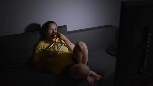 Wide shot of a young woman lying on a sofa and eating potato chips while watching a movie. The back of a television is partially visible in the frame, creating a relaxed home entertainment scene.