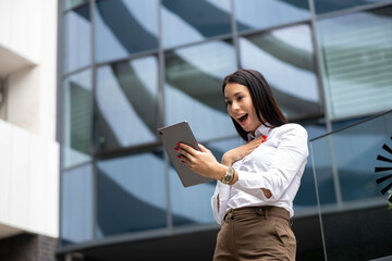Fototapeta premium Professional woman celebrating good news, receiving success on a tablet, happy with achievements in a modern business district