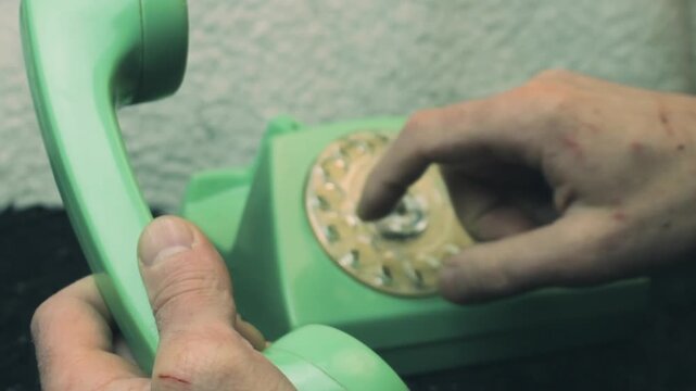 Close-up shot of a person's hands using a retro green rotary telephone. One hand holds the receiver while the finger dials a number. Soft focus highlights the nostalgic analog communication technology
