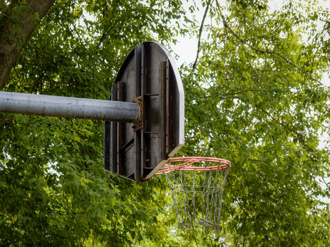 Rear side view of an outdoor steel basketball hoop backboard and chain net structure in a public park setting framed by green summer tree leaves