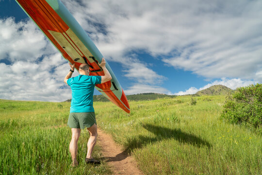 senior male paddler is carrying a long racing stand up paddleboard (SUP) uphill on a trail in early summer - Lory State Park, Colorado
