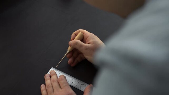 Craftsman using an awl and a metal ruler to mark a pattern on black genuine leather in his workshop