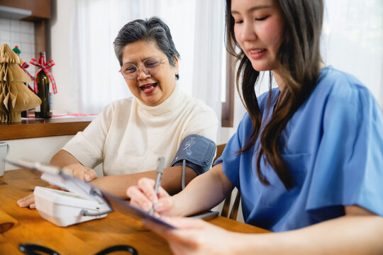 Nurse in blue scrubs measures blood pressure for an older patient and note. Patient smiles warmly while seated at wooden table. Blood pressure cuff is on patient arm in cozy home