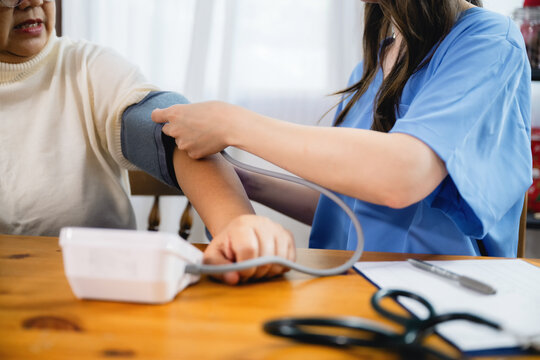 Healthcare professional measures blood pressure. She wears a blue uniform. The elderly woman relaxes her arm. Her expression is calm and cooperative. Medical tools sit on a wooden table