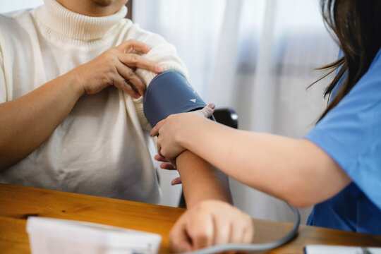 Healthcare worker checks blood pressure with cuff. Older adult sits at wooden table. Professional uses cuff on arm. She holds clipboard. Room has soft lighting.