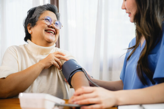 Woman smiles while having her blood pressure measured. She wears a light sweater, looks relaxed. Nurse in blue scrubs focuses on the task. They sit at a wooden table, near a window