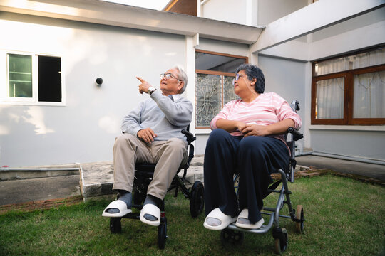 Elderly retired husband wife on wheelchairs looking up garden sharing moment together outside their home. old man points upward, old woman listens attentively beside him. Their connection feels warm