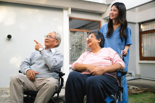 Older man points while seated in a wheelchair. An older woman smiles beside him in another wheelchair. Young woman in scrubs stand their side with warmth. Share moment senior couple.