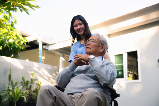 Elderly man in wheelchair looking worried, elderly man wears gray sweater and beige pants. Young caregiver stands behind him, smiling. Her hand rests on his shoulder. Old man looks calm and content