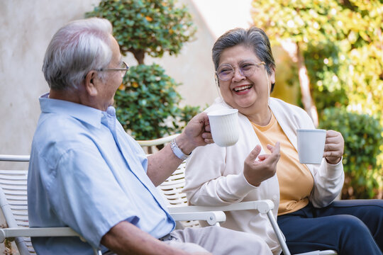 Elderly couple sits on white garden chairs. They hold white tea cups, smiling at each other. Lush green bushes and grass surround them. Their affectionate moment feels warm and peaceful
