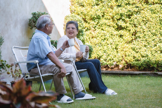 Elderly couple sits on garden chairs. They hold white teacups. Their smiles show quiet happiness. Green grass and bushes fill background. Stone wall frames the scene, Wide angle view with copy space