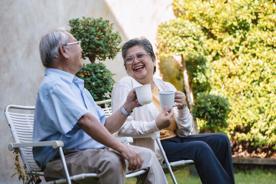 Elderly couple sits on white garden chairs. They hold white tea cups, laughing each other. Lush green bushes and grass surround them. Their affectionate moment feels warm and peaceful